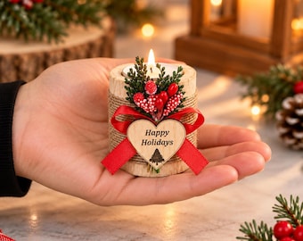 Może przedstawiać: Mały cylindryczny świecznik z drewna owinięty sznurkiem jutowym, z zapaloną świeczką typu tealight. Ozdobiony jest zielonymi gałązkami sosny, czerwonymi jagodami, czerwoną wstążką oraz drewnianym sercem z napisem Happy Holidays i drzewkiem.