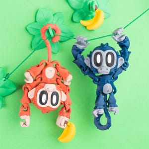 May include: Two toy monkeys, one orange and one blue, hang from a green string against a green background. Each monkey holds a yellow banana. The monkeys have large, round eyes and the number "00" on their chests. Green leaf-shaped decorations are in the background.