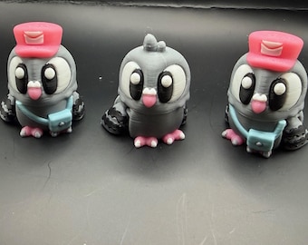 May include: Three 3D-printed pigeon figurines. Each gray pigeon has a pink hat, a blue messenger bag, and pink feet. The pigeons have large white eyes and are sticking out their tongues. The figurines are on a dark surface.