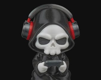 May include: A figurine of a skull-faced figure in a black hooded robe, wearing headphones with red accents. The figure is holding a game controller. The figurine is a unique decorative item.