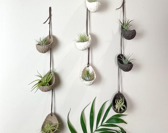 May include: Three hanging planters with air plants. The planters are made of ceramic and are in different colors and shapes. The planters are hanging from brown leather cords.