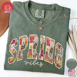 May include: Moss green t-shirt with the word "SPRING" in a patchwork floral design and the word "vibes" in cursive. A silver necklace with a pendant rests on the shirt. The shirt is size S-P-CH.