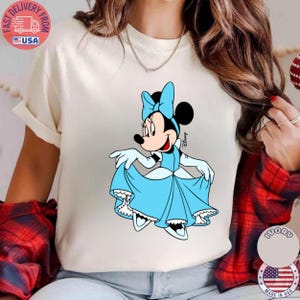 May include: Cream-colored t-shirt featuring a cartoon illustration of Minnie Mouse in a light blue dress and bow. The Disney character is smiling and holding up her dress. The shirt has a round neckline.