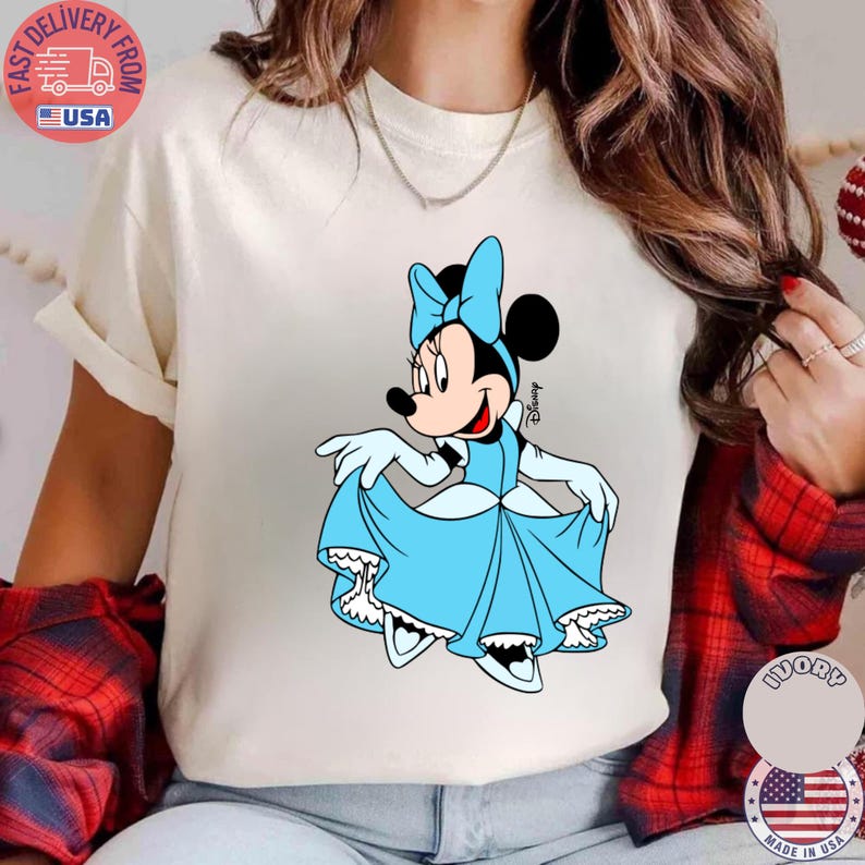 May include: Cream-colored t-shirt featuring a cartoon illustration of Minnie Mouse in a light blue dress and bow. The Disney character is smiling and holding up her dress. The shirt has a round neckline.