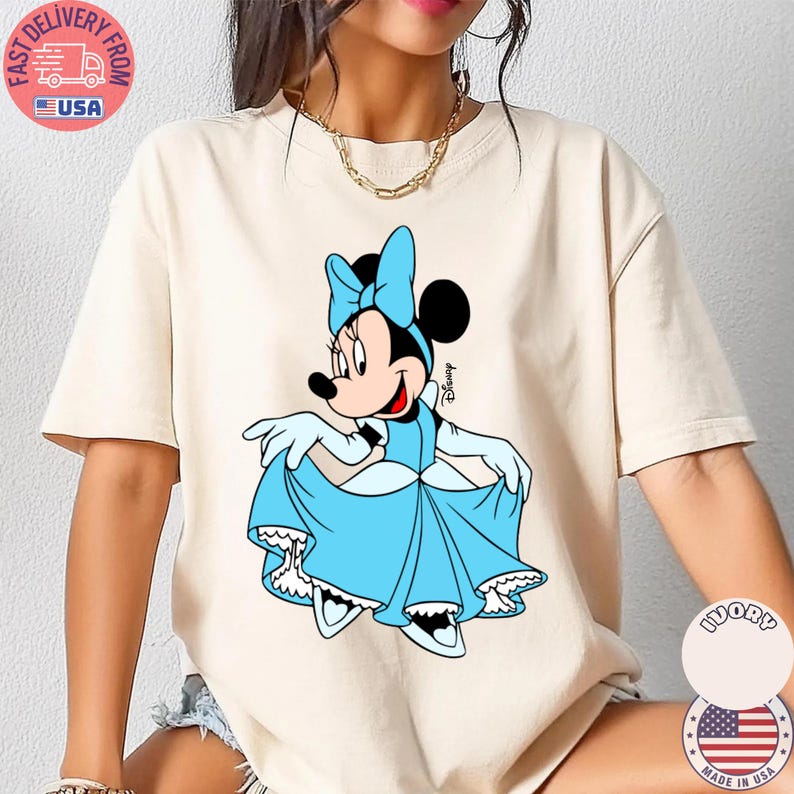 May include: Cream-colored t-shirt featuring a cartoon Minnie Mouse in a light blue dress and bow. The Disney character is smiling and holding up her dress. The shirt has a round neck and short sleeves.