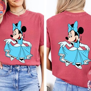 May include: A red-pink t-shirt featuring a Minnie Mouse graphic in a light blue dress and bow. The front and back of the shirt display the same design. The shirt has short sleeves and a relaxed fit. The shirt has a round neck.