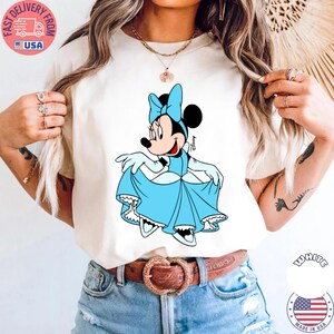 May include: Cream-colored t-shirt featuring a cartoon Minnie Mouse in a light blue dress and bow. The Disney character is smiling and holding up her dress. The shirt is paired with blue jeans and a brown belt.