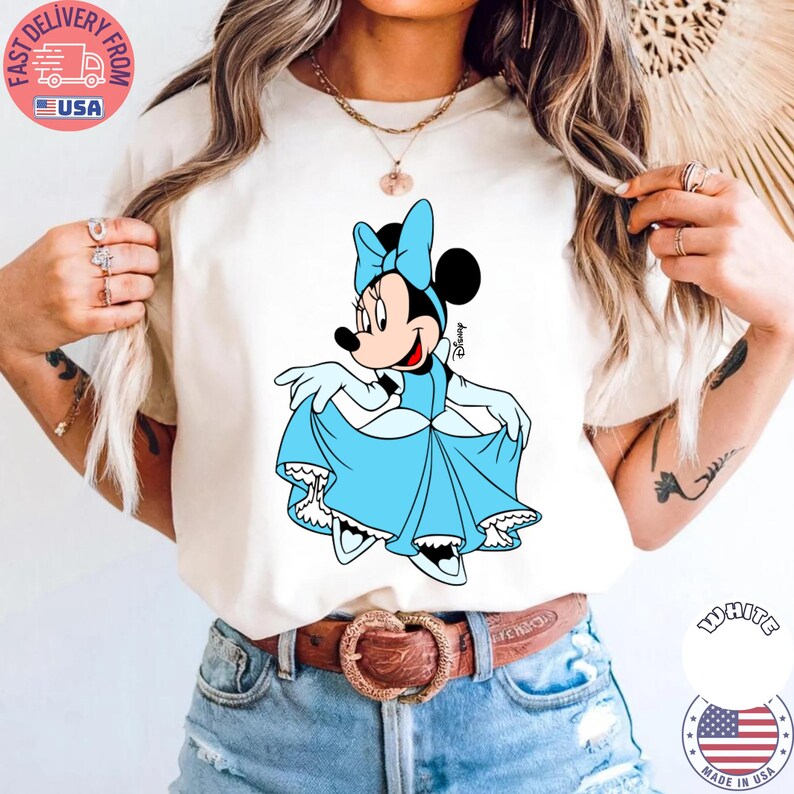 May include: Cream-colored t-shirt featuring a cartoon Minnie Mouse in a light blue dress and bow. The Disney character is smiling and holding up her dress. The shirt is paired with blue jeans and a brown belt.