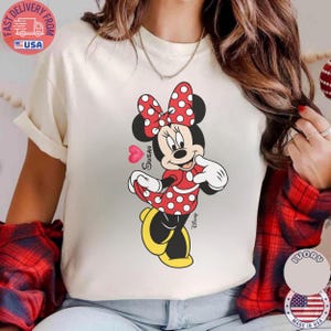 May include: Cream-coloured t-shirt with a Minnie Mouse graphic. Minnie is shown in a red dress with white polka dots, a matching bow, and yellow shoes. The name "Susan" is written on the graphic.