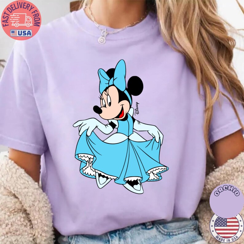 May include: A light purple t-shirt featuring a cartoon Minnie Mouse in a blue dress and bow. The Disney character is smiling and holding up her dress. The shirt has a crew neck and short sleeves.