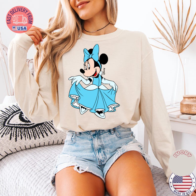 May include: Ivory-colored long-sleeve sweatshirt featuring a Minnie Mouse graphic in a light blue dress and bow. The Disney character is holding up her dress. The sweatshirt is paired with denim shorts.