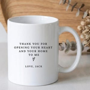 May include: A white ceramic mug featuring black printed text that reads: THANK YOU FOR OPENING YOUR HEART AND YOUR HOME TO ME, followed by a small hand-drawn heart symbol and the signature LOVE, JACK. The mug has a standard handle and cylindrical body.
