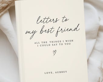 May include: A cream-coloured journal with the text "letters to my best friend" in elegant script. Below, it reads "ALL THE THINGS I WISH I COULD SAY TO YOU" with a heart symbol and "LOVE, AUBREY" at the bottom. The journal is on a white fabric.