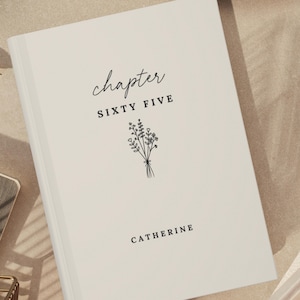 May include: A light beige hardcover book featuring the text "chapter SIXTY FIVE" in a mix of script and serif fonts, a black line art illustration of a small wildflower bouquet, and the name "CATHERINE" printed in black at the bottom.