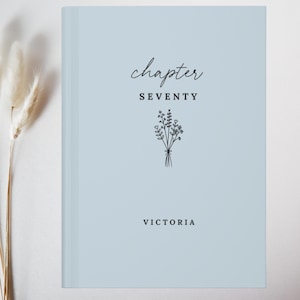May include: A light blue book with a hard cover featuring the text "chapter SEVENTY" and "VICTORIA" in black ink. A simple black line drawing of a small bouquet of wildflowers is centred between the text elements on the front cover.
