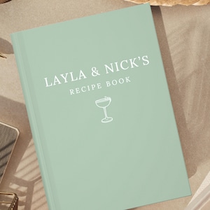 May include: A pale green recipe book with the text "LAYLA & NICK'S RECIPE BOOK" in white. A white line drawing of a cocktail glass is below the text. The book is on a neutral surface.
