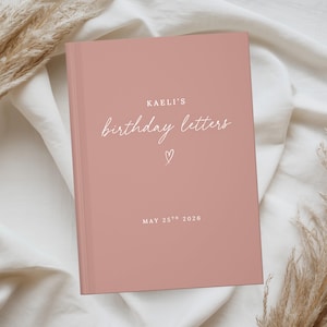 May include: A dusty rose hardcover book with white text that reads KAELI'S birthday letters, a small hand-drawn heart icon, and the date MAY 25TH 2026 printed on the front cover. The book has a smooth, matte finish and a clean, simple design.