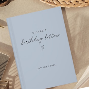 May include: A light blue hardcover book with black text that reads OLIVER'S birthday letters, a small hand-drawn heart symbol, and the date 21ST JUNE 2026 printed on the front cover. The book has a smooth, matte finish and a clean, simple design.
