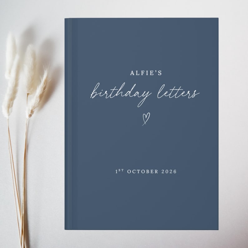 May include: A dark blue hardcover book with white text that reads ALFIE'S birthday letters, a small hand-drawn heart, and the date 1ST OCTOBER 2026. The book cover features a clean, solid matte finish with centered typography and a simple heart icon.
