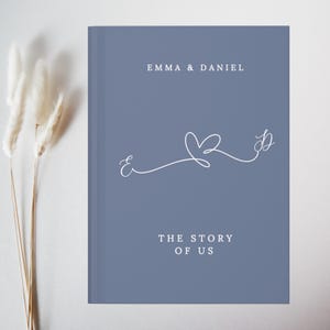 May include: A blue hardback book with the text "EMMA & DANIEL" at the top and "THE STORY OF US" at the bottom. A white line drawing of a heart and the letters "E" and "D" are in the centre.