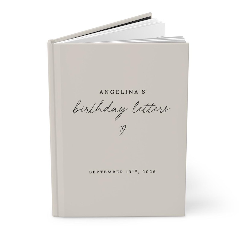 May include: A light beige hardcover book with white pages visible at the top. The cover features black text reading ANGELINA'S birthday letters, a small hand-drawn heart icon, and the date SEPTEMBER 19TH, 2026 printed in a clean, minimalist font.