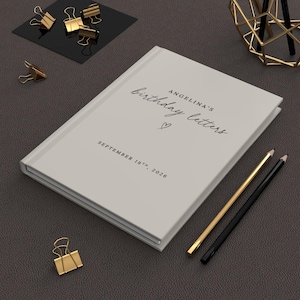 May include: A light gray hardcover book with the text "ANGELINA'S birthday letters" and "SEPTEMBER 19TH, 2026" printed on the front cover, accompanied by a small heart icon, positioned alongside gold-toned binder clips and two pencils.