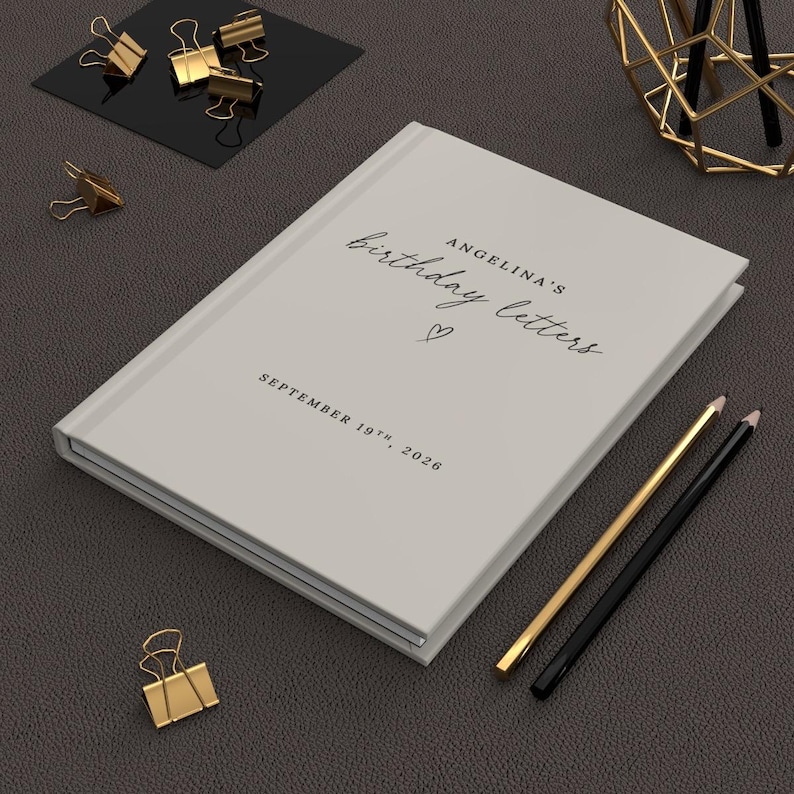 May include: A light gray hardcover book with the text "ANGELINA'S birthday letters" and "SEPTEMBER 19TH, 2026" printed on the front cover, accompanied by a small heart icon, positioned alongside gold-toned binder clips and two pencils.
