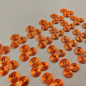 Pode incluir: Close-up de inúmeras espirais de arame cor de cobre, cada uma composta por três bobinas circulares conectadas. As espirais estão espalhadas sobre uma superfície cinza claro, criando um padrão de formas repetidas. O arame tem um acabamento metálico brilhante.