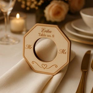 Könnte beinhalten: Ein Serviettenring aus Holz mit geometrischem Design. Der Ring hat eine kreisförmige Öffnung und ist mit dem Namen "Erika", der Tischnummer 8 und den Titeln "Mr" und "Ms" graviert. Ein Herz und dekorative Wirbel sind ebenfalls eingraviert.