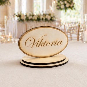 Può includere: Segnaposto in legno con il nome Viktoria. Il biglietto ovale è marrone chiaro con il nome "Viktoria" inciso in corsivo. Si erge su una base rotonda a due livelli con bordi neri, adatto per matrimoni o eventi.