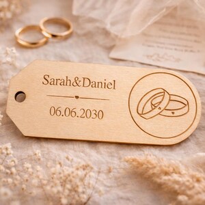 Könnte beinhalten: Ein hölzernes Hochzeitsanhänger mit der Gravur "Sarah & Daniel" und dem Datum "06.06.2030". Der Anhänger zeigt ein kreisförmiges Design mit zwei ineinandergreifenden Ringen. Zwei goldene Eheringe sind im Hintergrund, zusammen mit Trockenblumen.