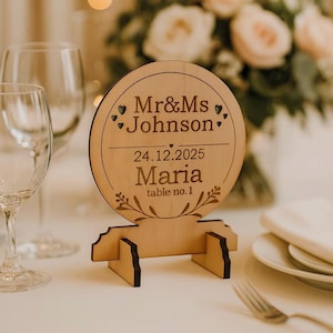 May include: A wooden table number sign with the text "Mr&Mrs Johnson" and "24.12.2025 Maria table no.1". The sign is round with a decorative floral design and is supported by a wooden stand. The background includes glasses and flowers.