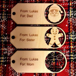 May include: Three wooden gift tags with laser-cut festive designs. Each tag features a circular cutout with a Santa, gingerbread man, and bell. Text on the tags reads "From: Lukas" and "For: Dad", "For: Sister", and "For: Mom".