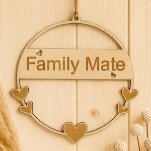 Könnte beinhalten: Runde Wanddekoration aus Holz mit dem Schriftzug "Family Mate". Das Design umfasst herzförmige Ausschnitte und eine Juteschnur zum Aufhängen. Der Hintergrund ist eine helle Holzplatte.
