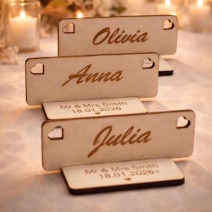 May include: Wooden place cards with names Olivia, Anna, and Julia engraved in a cursive font. Each card has a heart-shaped cutout and sits in a matching base. The bases read "Mr & Mrs Smith 18.01.2026".