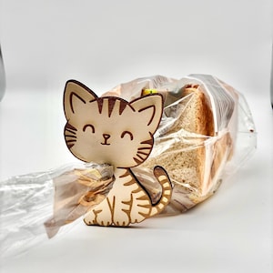 Könnte beinhalten: Ein brotförmiger Clip in Katzenform aus Holz mit einem lächelnden Gesicht und Tigerstreifen. Der Clip ist hellbraun und beige und hält einen durchsichtigen Plastikbeutel mit geschnittenem Brot. Der Hintergrund ist weiß.