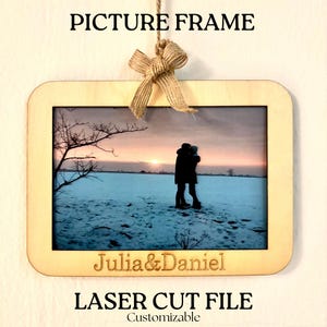 Könnte beinhalten: Ein Holz-Bilderrahmen mit abgerundeten Ecken, der ein Foto eines Paares in einer verschneiten Landschaft bei Sonnenuntergang zeigt. Der Rahmen ist mit einer Jute-Schleife verziert und die Namen "Julia & Daniel" sind eingraviert. Der Text oben lautet "PICTURE FRAME" und "LASER CUT FILE".