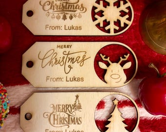 Peut inclure: Trois étiquettes cadeaux de Noël en bois avec des motifs découpés au laser. Chaque étiquette porte l'inscription "Merry Christmas" et "From: Lukas". L'une présente un flocon de neige, une autre un renne et la dernière un sapin de Noël.