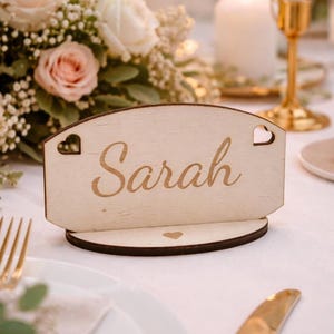 May include: A wooden place card with the name "Sarah" engraved in a cursive font. The card has heart-shaped cutouts and sits on a round base. The background includes flowers and a candle.