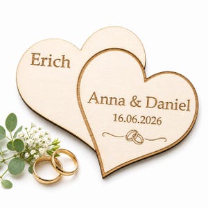 Könnte beinhalten: Zwei herzförmige Hochzeitsdekorationen aus Holz. Ein Herz ist mit dem Namen "Erich" graviert, das andere mit "Anna & Daniel" und dem Datum "16.06.2026". Goldene Eheringe und Grünzeug sind im Vordergrund.