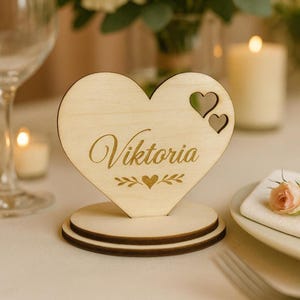 Könnte beinhalten: Ein herzförmiger Platzkartenhalter aus Holz mit dem Namen "Viktoria" in Schreibschrift graviert. Das Herz hat zwei kleinere Herz-Ausschnitte und ruht auf einem runden Sockel. Dekorative Elemente sind ein Herz- und Blätterdesign. Der Halter ist aus hellem Holz gefertigt.