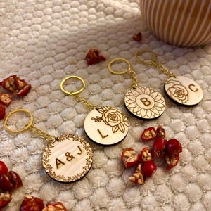 May include: Wooden keychains with gold-colored chains and key rings. The keychains feature engraved floral designs and initials. One keychain has the initials "A & J", another has the initial "L", and another has the initial "C". A sunflower design is also visible.