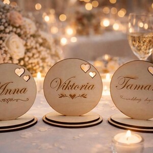 Puede incluir: Tarjetas de mesa de madera con los nombres Anna, Viktoria y Tama. Cada tarjeta redonda tiene un recorte de corazón y se asienta sobre una base de dos niveles. La tarjeta Viktoria tiene un diseño floral. Tema de boda.