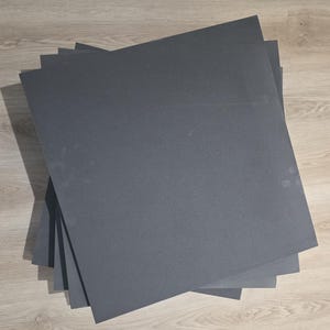 May include: A pile of dark grey rectangular sheets, possibly paper or card, arranged on a light wood-grain surface. The sheets are uniformly coloured and appear to be of a consistent size and thickness. The lighting creates shadows, adding depth to the image.