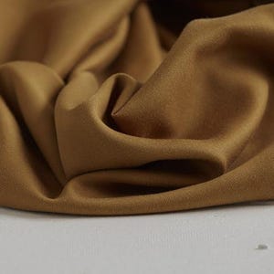 Könnte beinhalten: Nahaufnahme eines gefalteten Stoffes in einem warmen, goldbraunen Farbton. Der Stoff hat eine glatte Textur mit subtilen Falten und Schatten, die ein Gefühl von Tiefe und Weichheit erzeugen. Das Material scheint ein mittelschweres Textil zu sein.