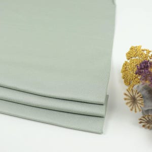 May include: A stack of three folded, light green fabric pieces. The fabric has a ribbed texture and is neatly arranged. Dried yellow and purple flowers are in the background.