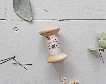 Pode incluir: Uma bobina de madeira com fita rosa pálido com um padrão floral em vermelho e azul marinho. A bobina é rodeada por alfinetes, folhas de eucalipto e pequenas flores brancas, tudo sobre um fundo branco.