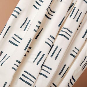 Könnte beinhalten: Nahaufnahme eines weißen Stoffes mit einem sich wiederholenden Muster aus dunkelblauen Strichen und horizontalen Linien. Der Stoff ist drapiert und zeigt Falten und Schatten. Das Design ist schlicht und modern, geeignet für Heimtextilien oder Kleidung.
