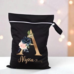 Puede incluir: Bolsa reutilizable negra con cremallera blanca y asa negra. La bolsa presenta la letra "A" dorada con un diseño floral en azul marino, rosa y verde. El nombre "Alyssa" está escrito en oro debajo.