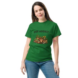May include: Green t-shirt featuring the words "POT ADDICT" above a graphic of potted plants. The illustration includes various plants and flowers in terracotta pots. The shirt is worn by a person.